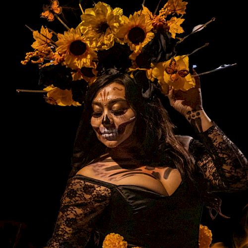 A person wearing a floral headpiece and skull face paint, dressed in a black lace outfit, posed dramatically against a dark background.