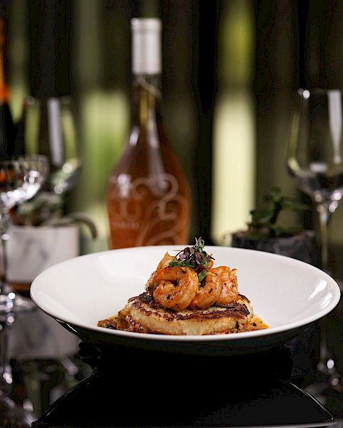 A plate of gourmet food is in focus, with shrimp on toast, surrounded by wine glasses and bottles in the background.