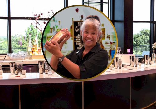 A bartender smiling while shaking a cocktail shaker is overlaid on an image of a well-lit, modern bar setup.